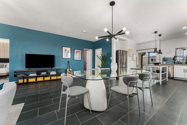 A modern living room with a glass dining table, grey chairs, a flat-screen TV, and a kitchen area with white cabinets and a stainless steel refrigerator.