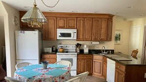 A compact kitchen with wooden cabinets, a white refrigerator, a microwave, a stove, and a sink. There is a dining table with chairs and a chandelier above.