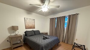 Un dormitorio con cama, mesita de noche, lámpara, ventilador de techo y ventana con cortinas.