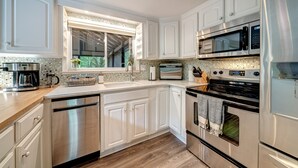 Una cocina moderna con armarios blancos, electrodomésticos de acero inoxidable, un amplio ventanal y piso de madera.