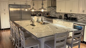 Cozinha moderna com uma ilha grande, armários brancos, eletrodomésticos de aço inoxidável e iluminação pendente.