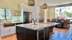 Een moderne keuken met een groot eiland, witte kasten, een zwarte koelkast en een eethoek met rieten stoelen.