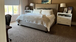 Une chambre à coucher comprenant un lit, deux tables de chevet, un ventilateur de plafond, une fenêtre avec des rideaux et un tableau accroché au mur.