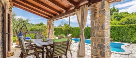 A covered outdoor seating area with a table and chairs, a hanging chair, and a stone pillar.