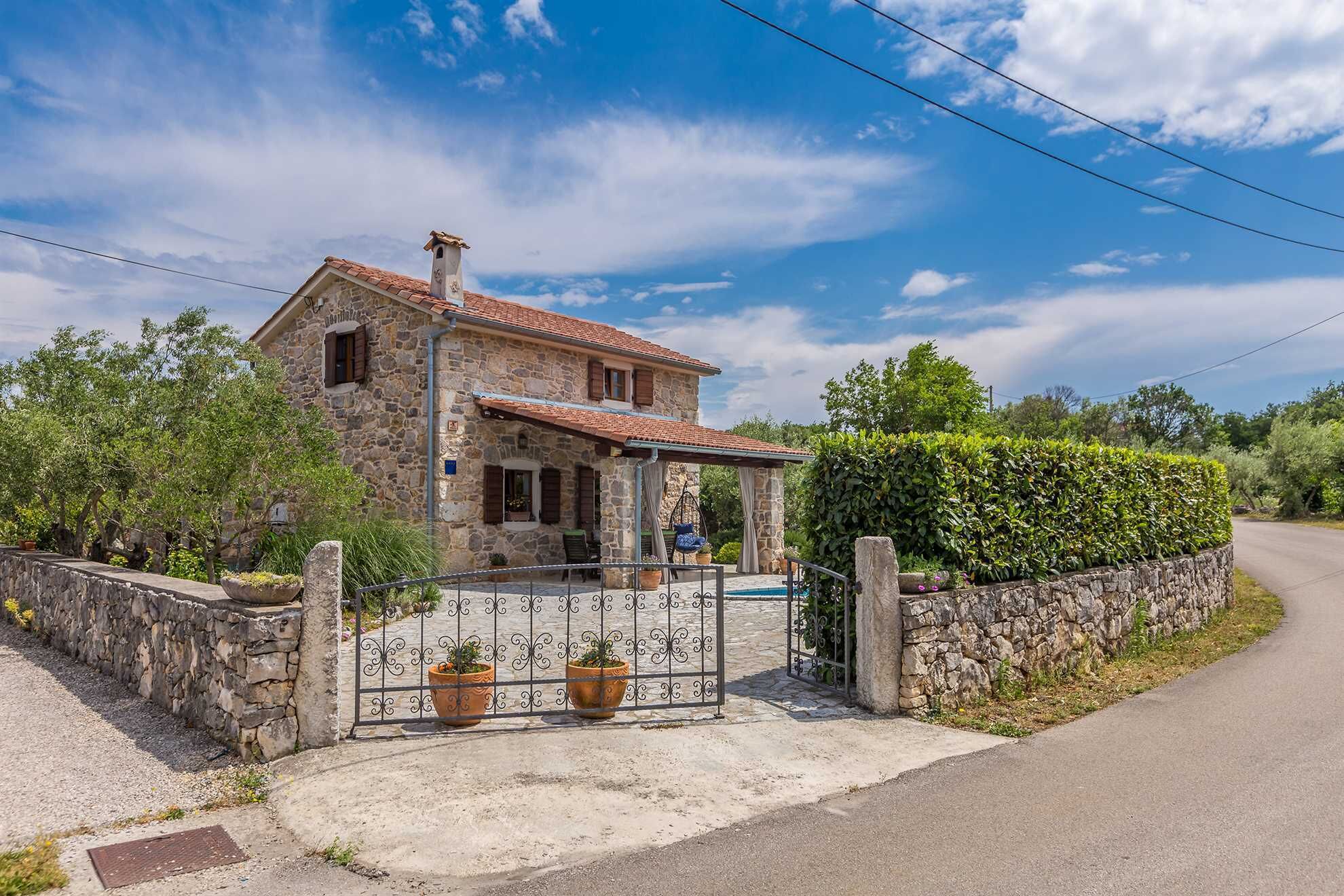 Una casa in pietra con tetto in tegole, un muro in pietra e un cancello metallico con piante in vaso.