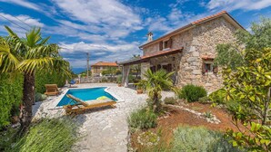 A stone villa with a swimming pool, surrounded by lush greenery and a clear blue sky.