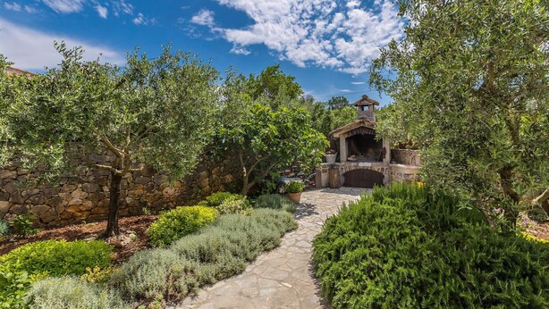 A stone pathway leading to a rustic stone structure with a chimney, surrounded by lush greenery and olive trees.