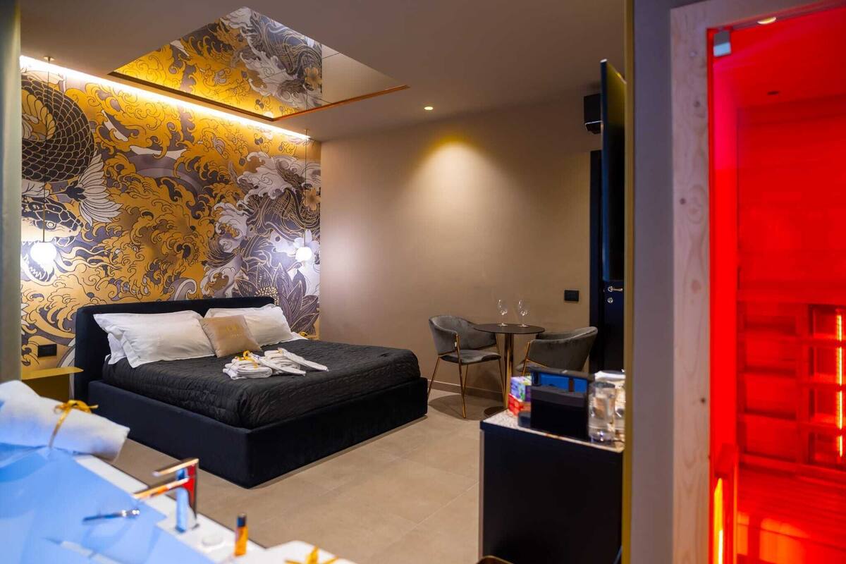 Quarto de hotel moderno com uma cama grande, uma escrivaninha com computador, uma mesinha com duas cadeiras e uma parede decorativa com arte tradicional.