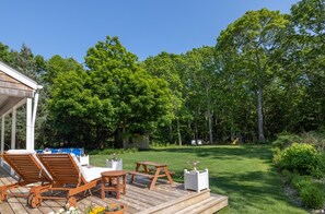 A wooden deck with lounge chairs and a table, surrounded by a lush green lawn and trees.