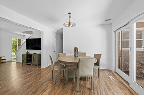 A dining area with a round table, six chairs, and a chandelier. There is a television and a cabinet in the background.