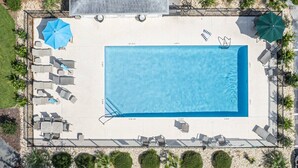 Une vue aérienne d’une piscine rectangulaire, bordée de chaises longues et de parasols, et entourée d’un espace paysager agrémenté de petits arbres et d’arbustes.