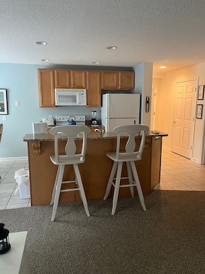 A compact kitchen with wooden cabinets, a microwave, a refrigerator, and a small island with two bar stools.