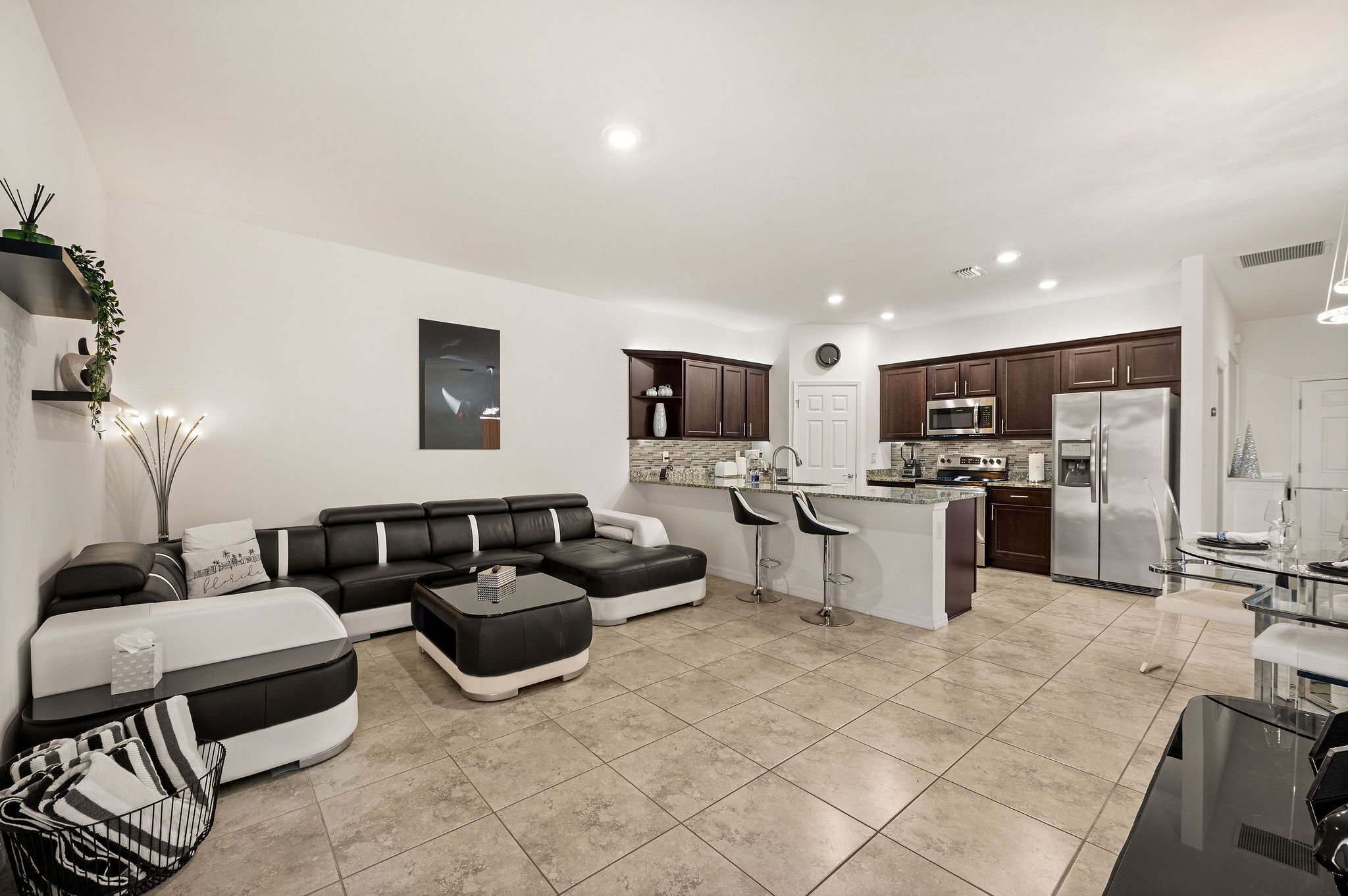 A modern living room with a sectional sofa, a glass dining table, and a kitchen area with dark wooden cabinets and stainless steel appliances.