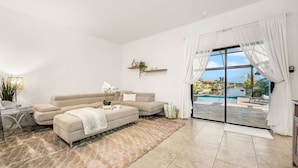 Ein modernes Wohnzimmer mit einem beigen Sofa, einem Glastisch und Blick auf einen Swimmingpool durch große Glasüren.