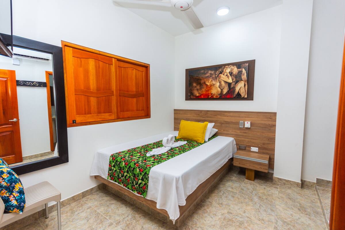 Une chambre d’hôtel avec un lit, une tête de lit en bois, un tableau au mur, un miroir et une fenêtre avec des volets en bois.
