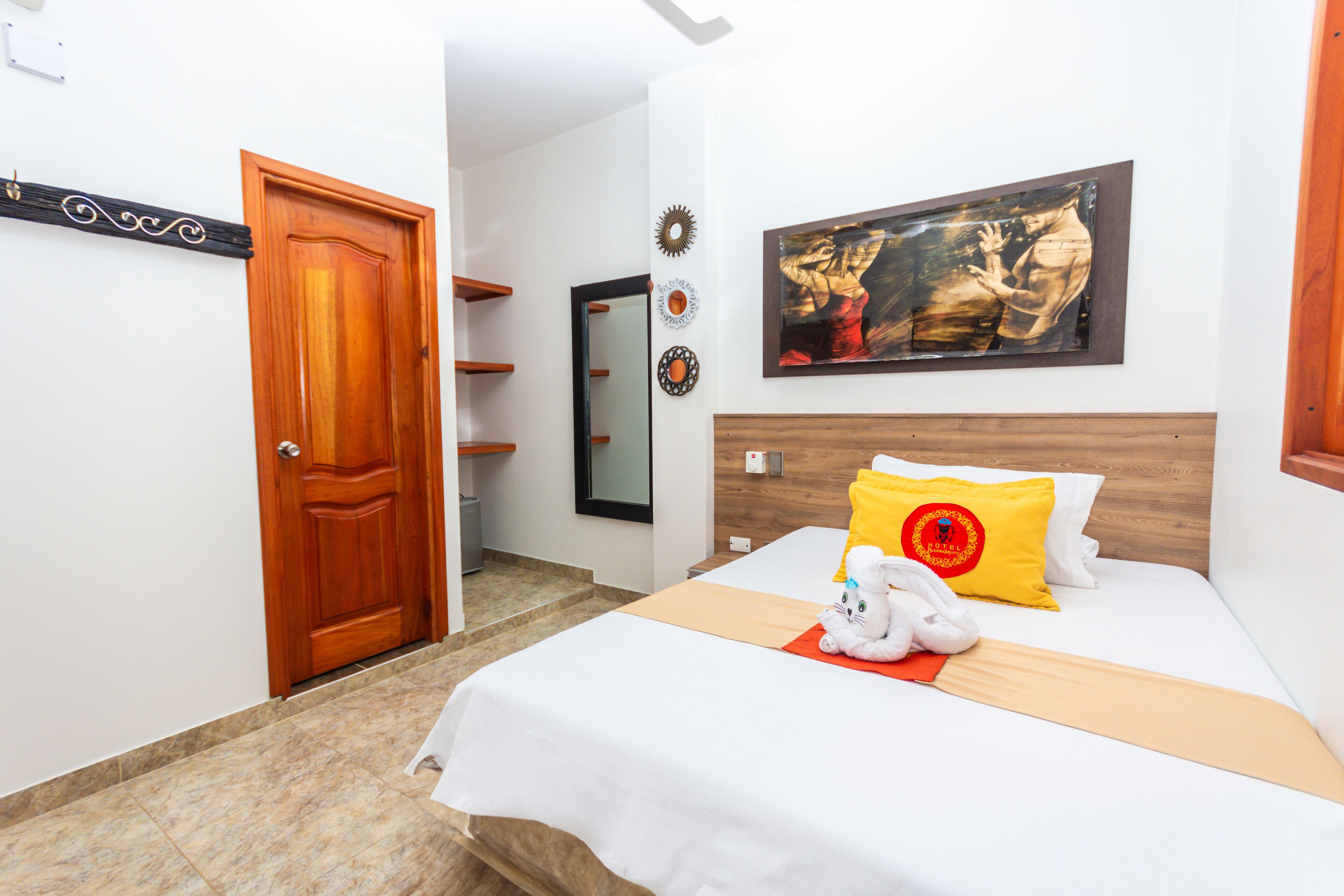 A hotel room with a wooden door, a bed with white bedding, a yellow pillow with a red emblem, a white stuffed toy, and a painting on the wall.