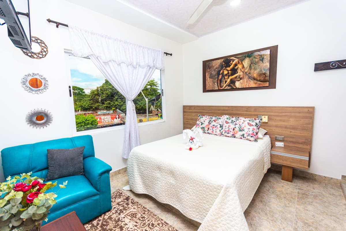 Une chambre à coucher avec un lit, un canapé bleu, une fenêtre avec des rideaux, un tableau au mur et une vue sur l’extérieur.
