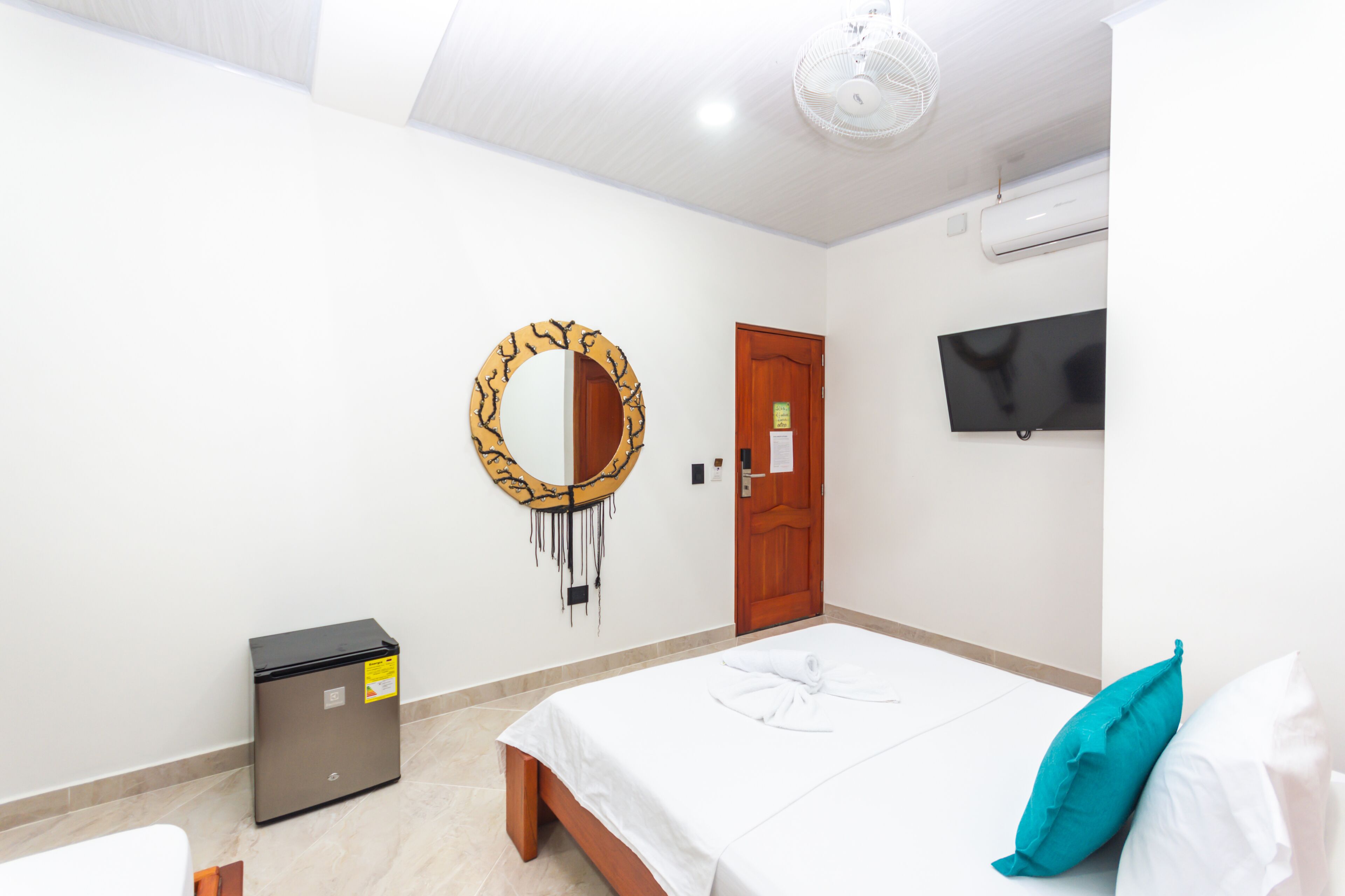 Ein Hotelzimmer mit Bett, kleinem Schreibtisch, Spiegel, Fernseher und Deckenventilator.