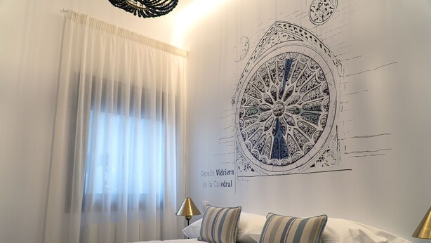 A bedroom with a large bed, two pillows, a wall mural, a window with sheer curtains, and a lamp on the nightstand. - CATEDRAL CERVANTES 5 COLLECTION (Toledo)