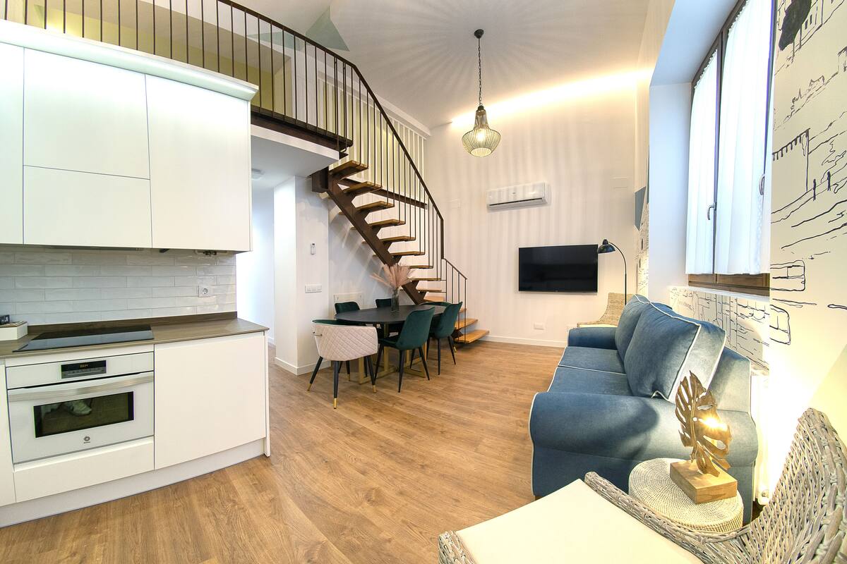 A modern kitchen with white cabinets, a built-in oven, and a dining area with a table and chairs. A staircase with a wooden railing leads to an upper level.