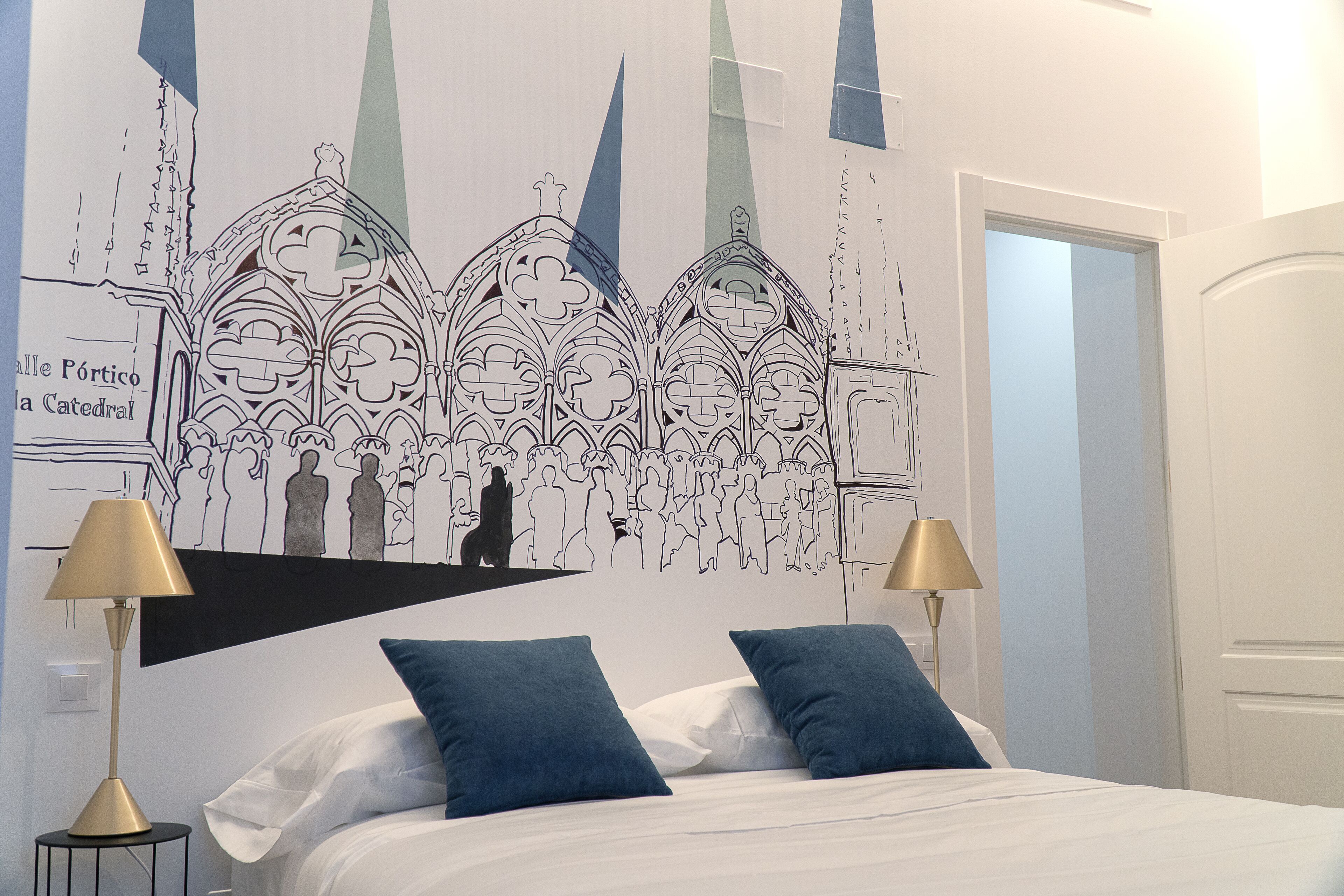 Un dormitorio con una cama, dos almohadas azules, dos lámparas de noche y un mural en la pared con un paisaje de una catedral.