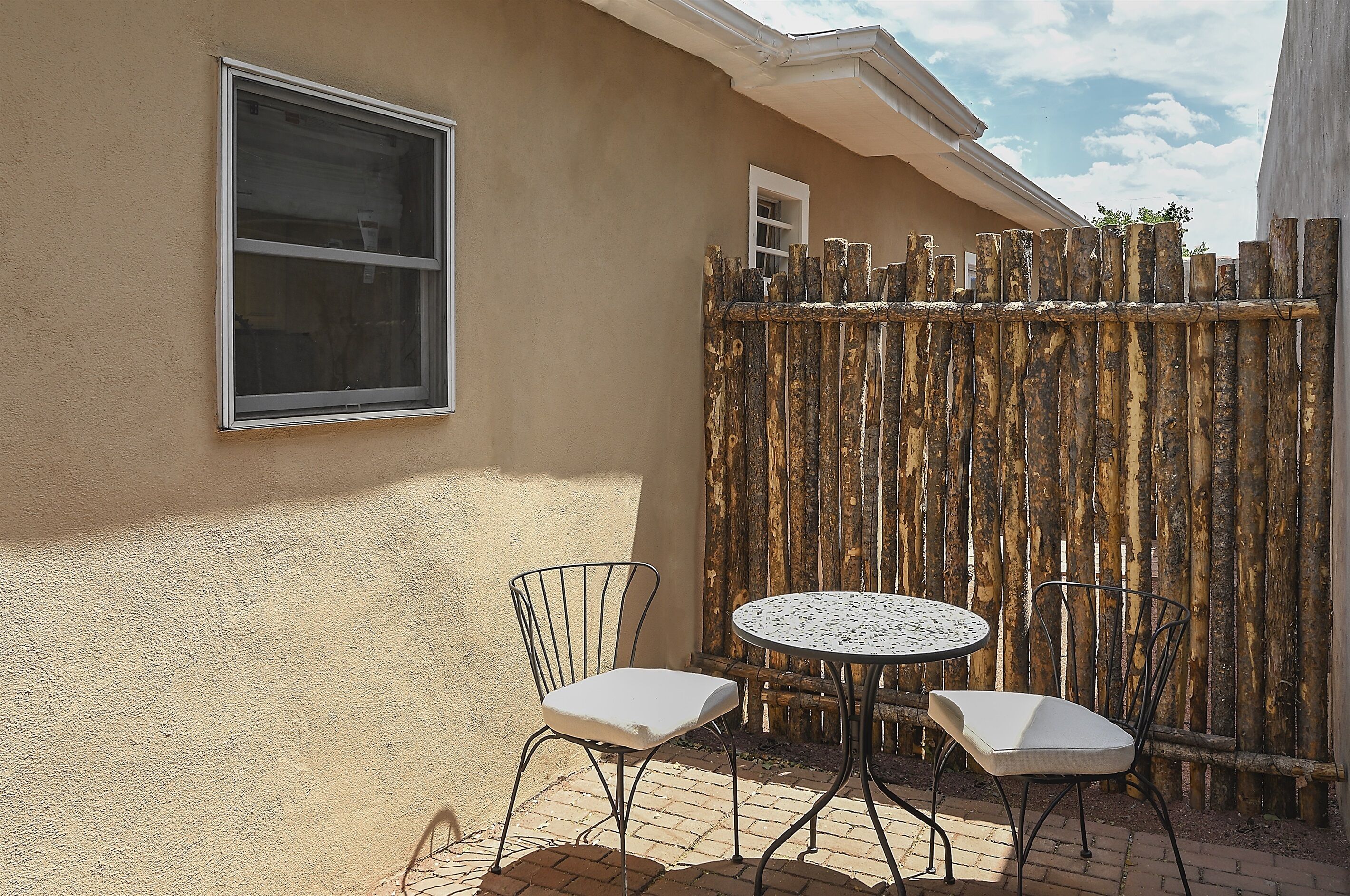 A patio with two white cushioned chairs and a small round table, a window, and a wooden fence.