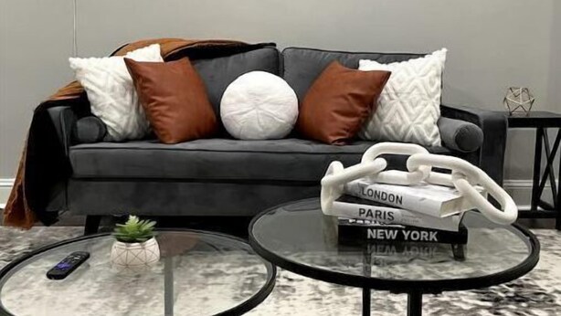 Ein modernes Wohnzimmer mit einer grauen Couch, mehreren Kissen, zwei Glastischen und einer Wanddekoration.