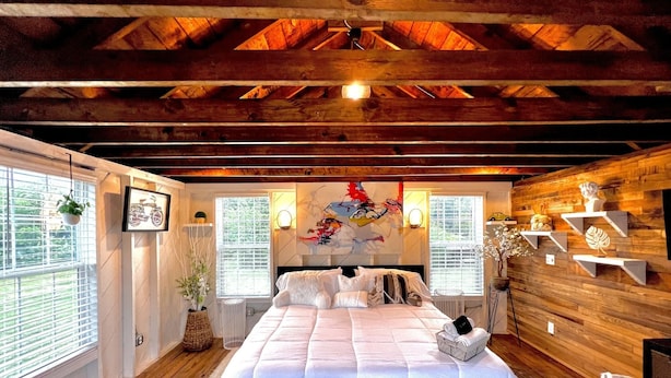 A spacious bedroom with a large bed, wooden beams on the ceiling, and a view of the outdoors through the windows. - Tiny House Hideaway with King sized Bed (Cape Charles)