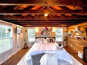 A spacious bedroom with a large bed, wooden beams on the ceiling, and a view of the outdoors through the windows. - Tiny House Hideaway with King sized Bed (Cape Charles)