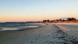 A beach with a long stretch of sand, a few scattered rocks, and a distant shoreline with buildings. - Tiny House Hideaway with King sized Bed (Cape Charles)