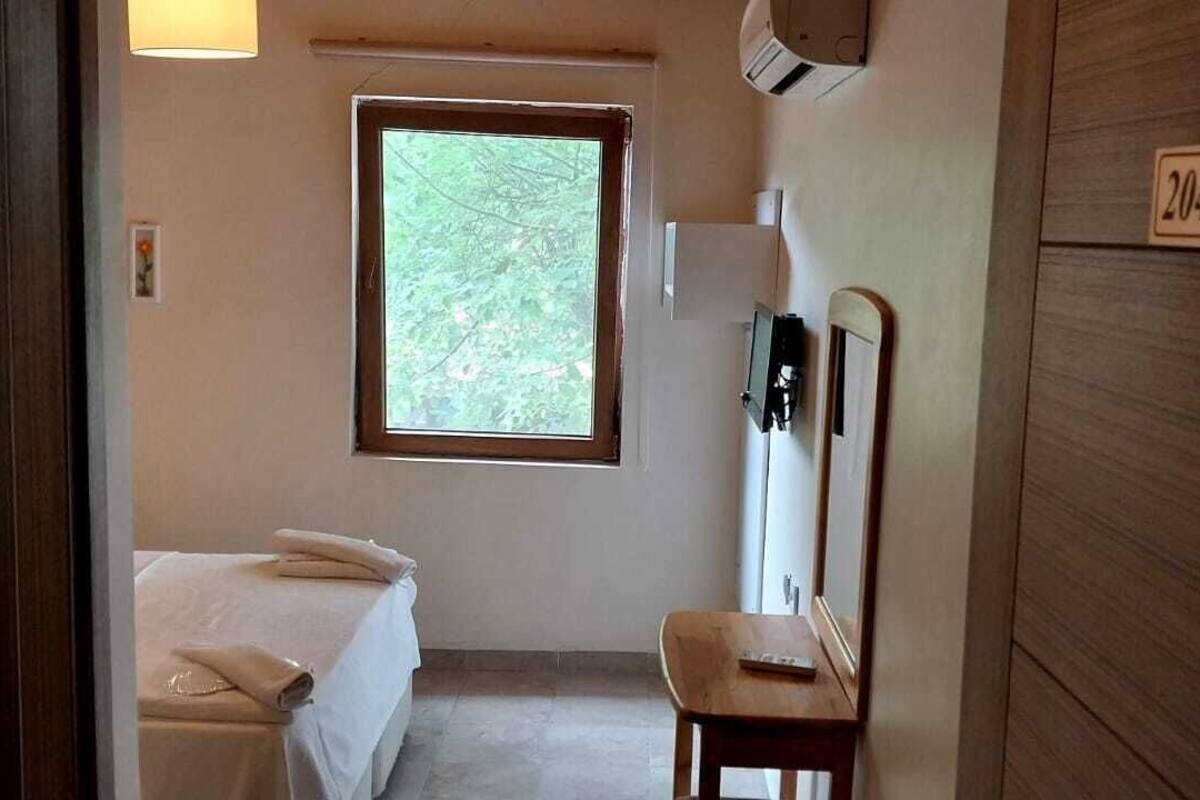 Une petite pièce agréable, dotée d’un lit, d’une fenêtre donnant sur un paysage verdoyant, d’un miroir et d’un panneau en bois portant un numéro.