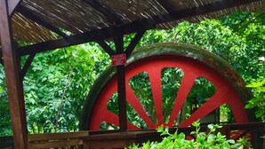 A wooden bench under a thatched roof structure, surrounded by lush greenery and a large red wheel. - Uyku Vadisi Otel (Milas)