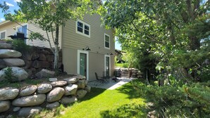 A two-story house with a stone retaining wall, a grassy yard, and a patio with outdoor furniture.