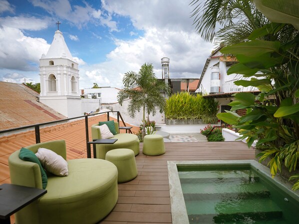 Eine Dachterrasse mit grünen Sitzgelegenheiten, einem kleinen Pool und Blick auf eine weiße Kirche mit Kreuz.