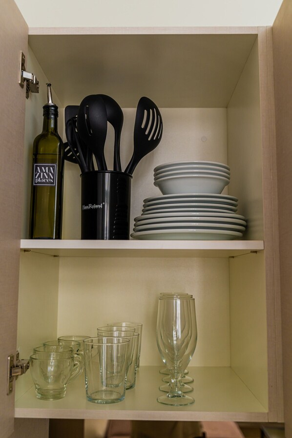 A kitchen cabinet with a wine bottle, black utensils, stacked plates, and clear drinking glasses.