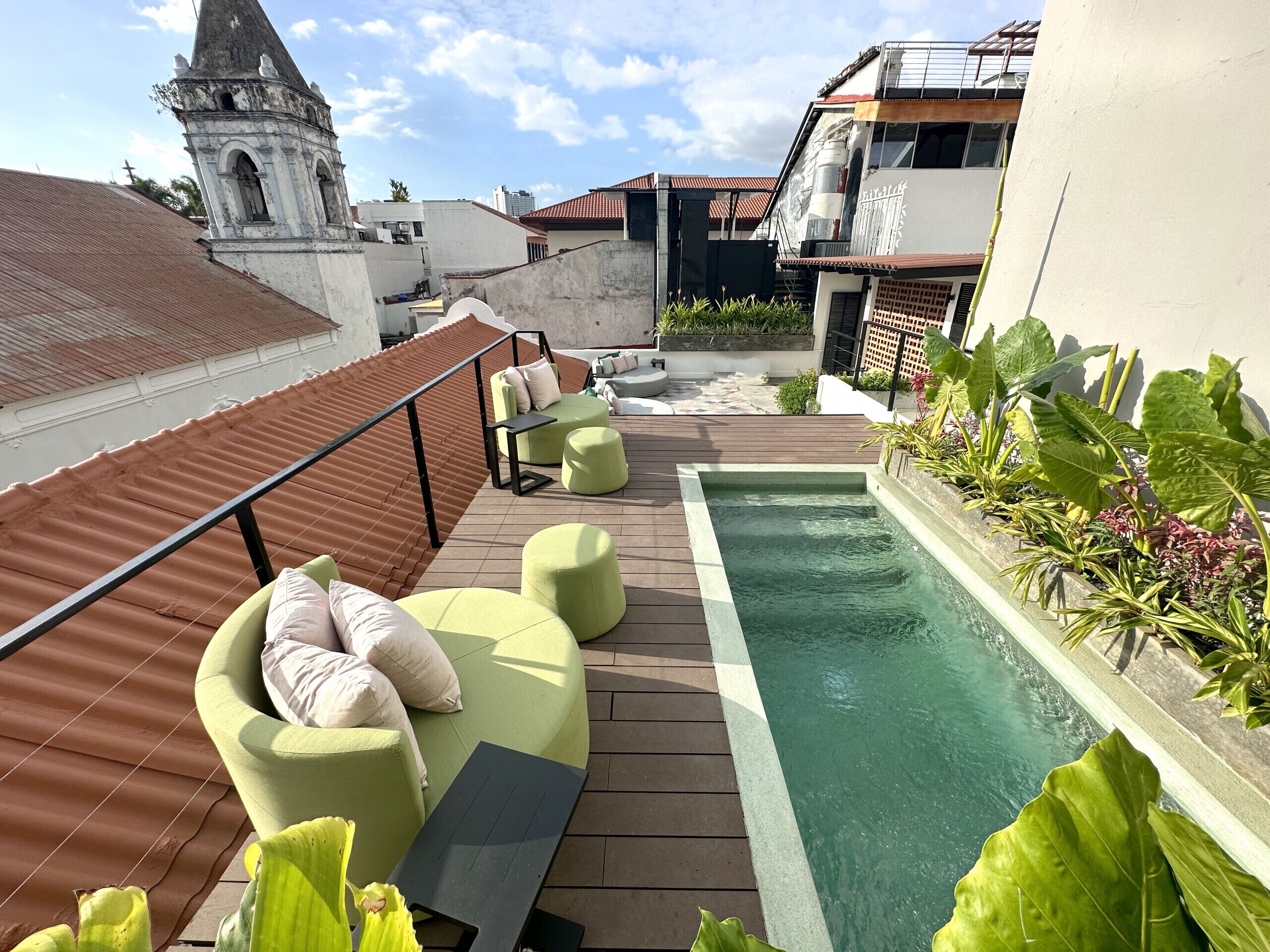 Una terraza en la azotea con piscina, asientos acolchados verdes y vistas a una torre histórica.