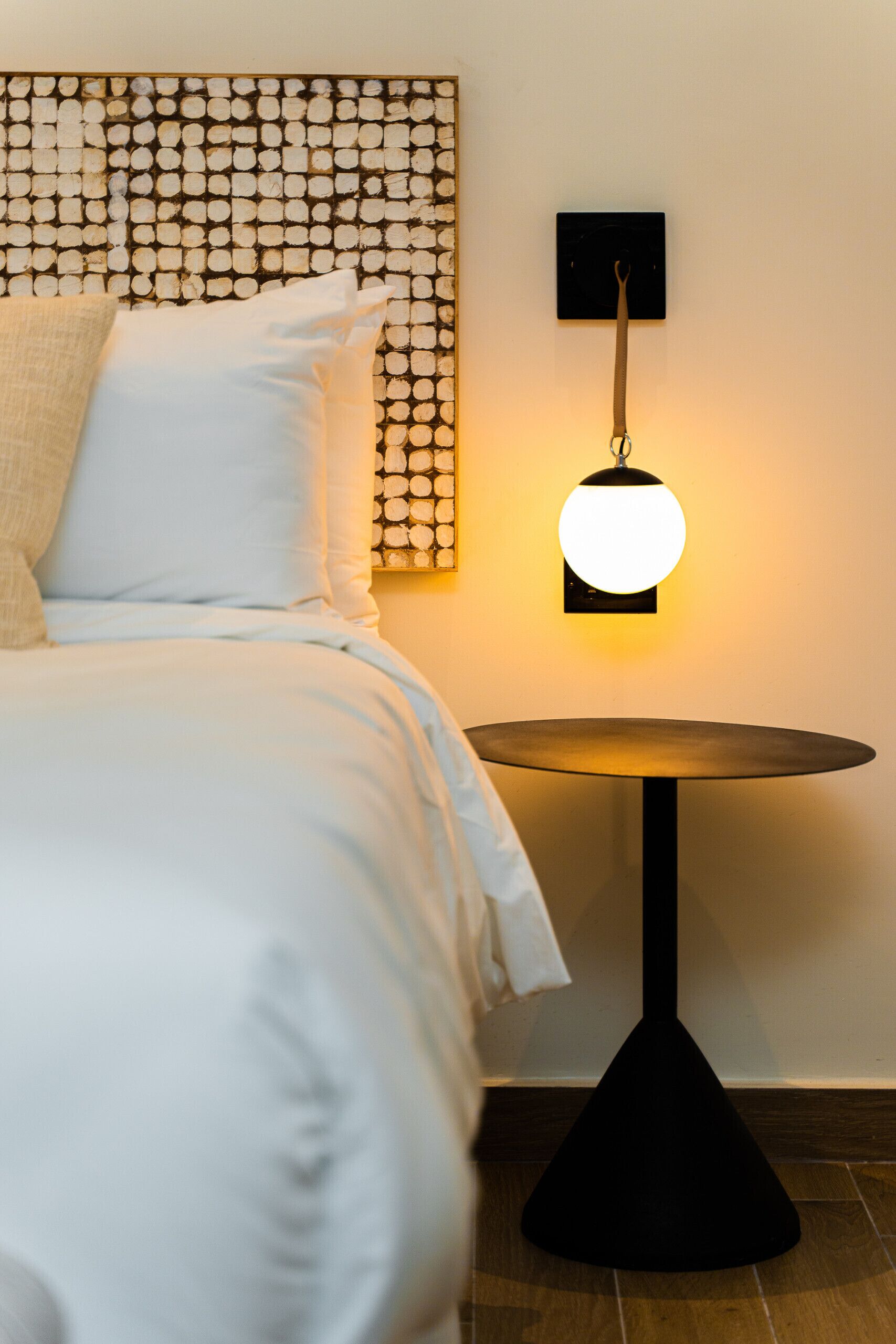 A neatly made bed with white bedding and a headboard with a circular pattern. A round table with a black base is positioned next to the bed, and a wall-mounted lamp with a spherical light fixture is visible.