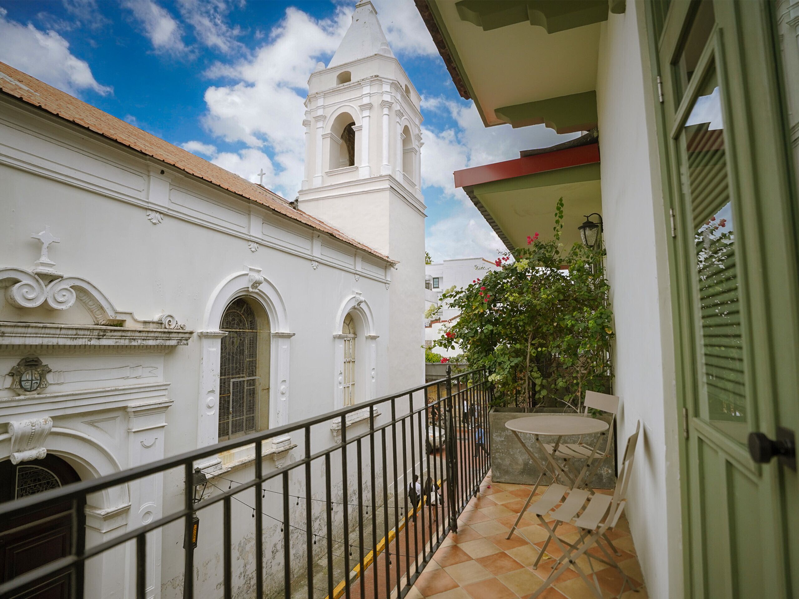 A balcony with a tiled floor, a metal railing, and a view of a white building with a bell tower.