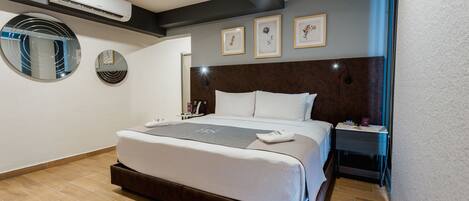 A modern hotel room with a large bed, a bedside table, a nightstand, a wall-mounted air conditioner, and framed artwork on the wall.