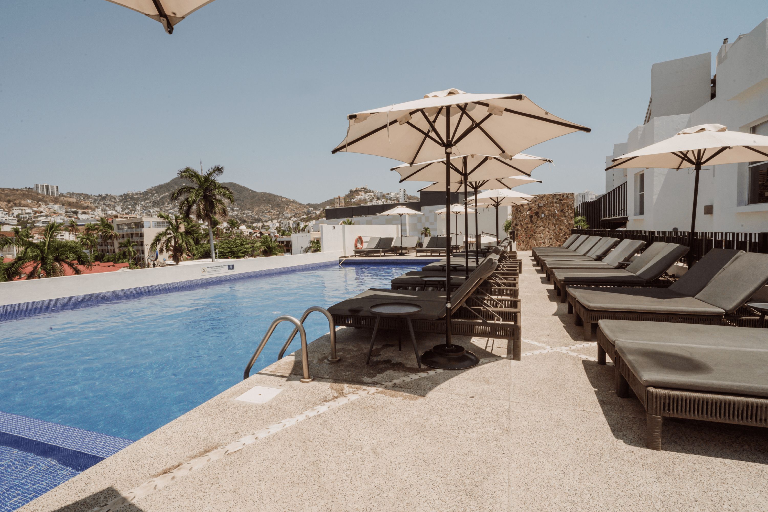 Zona de piscina con sillones de descanso y sombrillas, una piscina y vistas a un pueblo en la colina.
