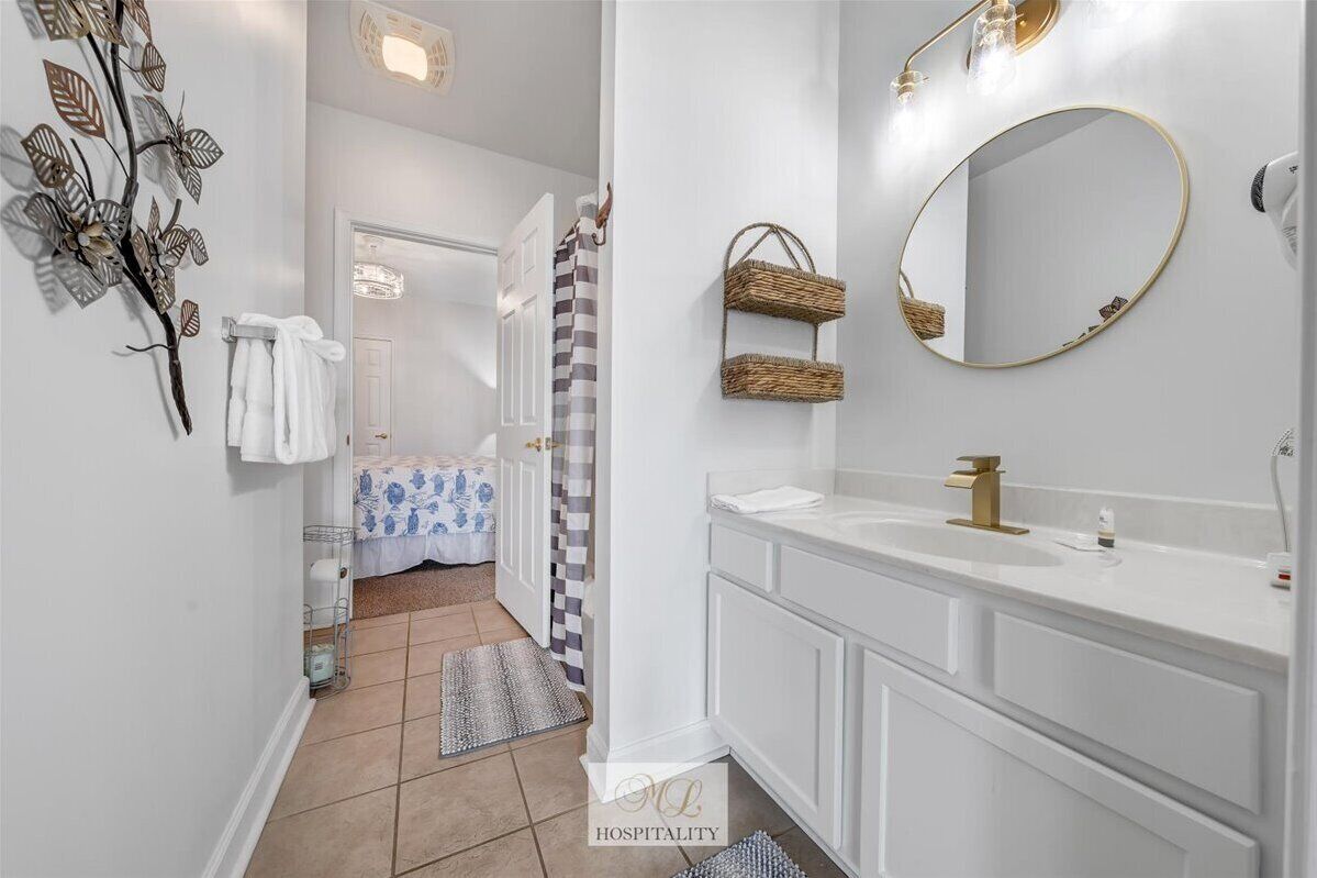 Une salle de bain moderne avec un meuble-lavabo blanc, des robinets dorés et un grand miroir. On y trouve des paniers en osier accrochés au mur et une branche décorative ornée de feuilles.