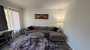 Un salon comprenant un canapé d’angle, une table basse en verre, un tapis à motifs, un tableau accroché au mur et une lampe.