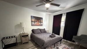 Une chambre à coucher comprenant un lit, une table de chevet, une lampe, un ventilateur de plafond, une fenêtre avec des rideaux et une chaise.