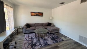 Un salon avec un canapé d’angle gris, une table basse en verre, un tapis à motifs, un bureau avec une chaise et un tableau accroché au mur.