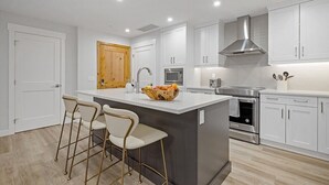 Uma cozinha moderna com armários brancos, uma ilha central com banquetas, eletrodomésticos de aço inoxidável e uma porta de madeira.