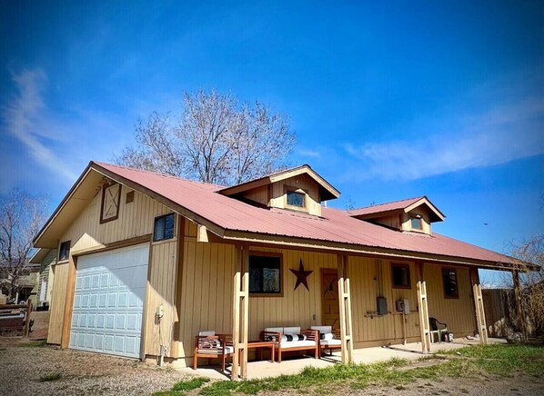 Une maison en bois à deux étages, avec un toit rouge, un garage et une véranda dotée de sièges pour l’extérieur.