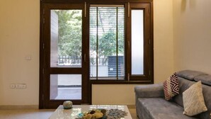A living room with a sofa, a coffee table with decorative items, and a view through a window with blinds. - Cozy Getaway: Clean and modern!!!    Breakfast Included !! (Gurugram)