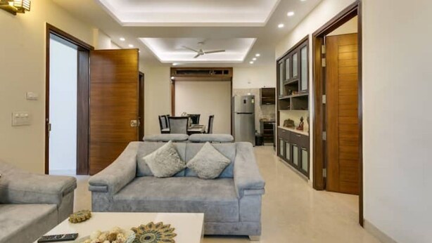 A modern living room with a grey sofa, a coffee table with decorative items, and a dining area in the background. - Cozy Getaway: Clean and modern!!!    Breakfast Included !! (Gurugram)