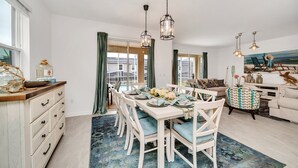 Een eetgedeelte met een witte tafel gedekt voor zes personen, omringd door witte stoelen met blauwe kussens. Boven de tafel hangen hanglampen.