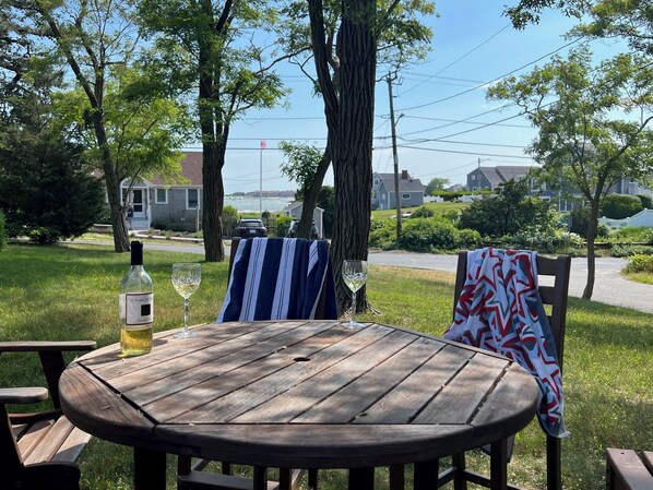 A wooden picnic table with a bottle of Charles wine and a glass, set outdoors with a view of houses and a body of water in the background.